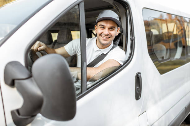 Portrait of a cheerful delivery driver in uniform looking out the window of the white cargo van vahicle, delivering goods by car
